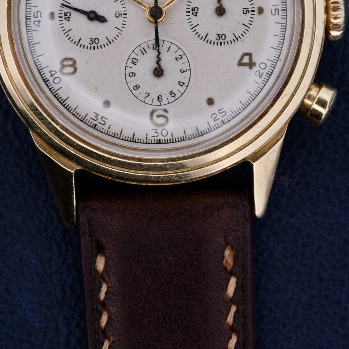 Close-up of a Vintage Movado For Tiffany & Co 14k Gold Chronograph M95 wristwatch with an odd dot dial, white face, and brown leather strap. It rests on a dark blue surface, exuding the timeless sophistication typical of Tiffanys esteemed collections.