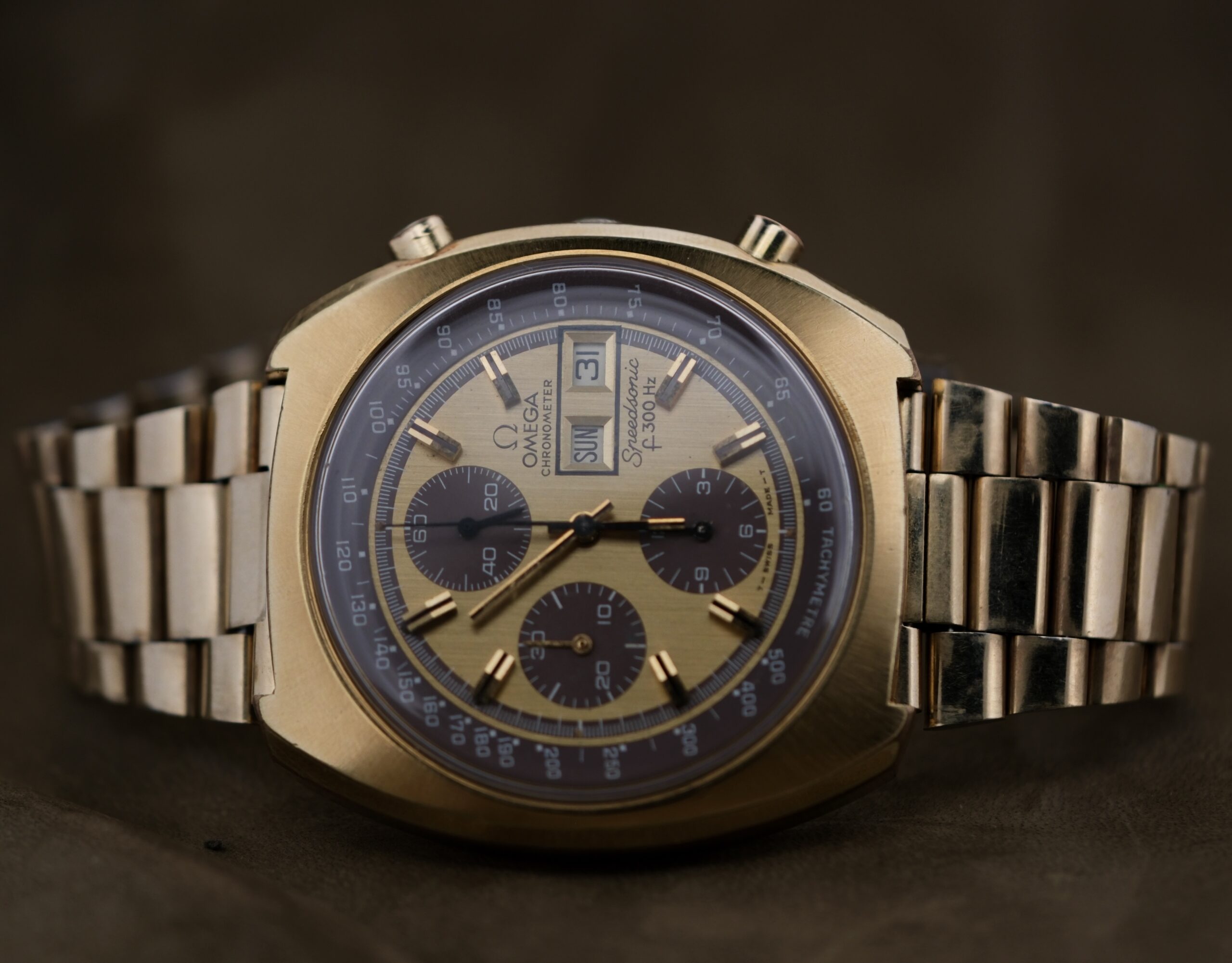 The 1975 Omega Speedsonic f 300Hz Chronograph Ref. 188.0002 features a gold detailed face with three sub-dials and a date window, paired with a metallic bracelet, set elegantly against a dark background.