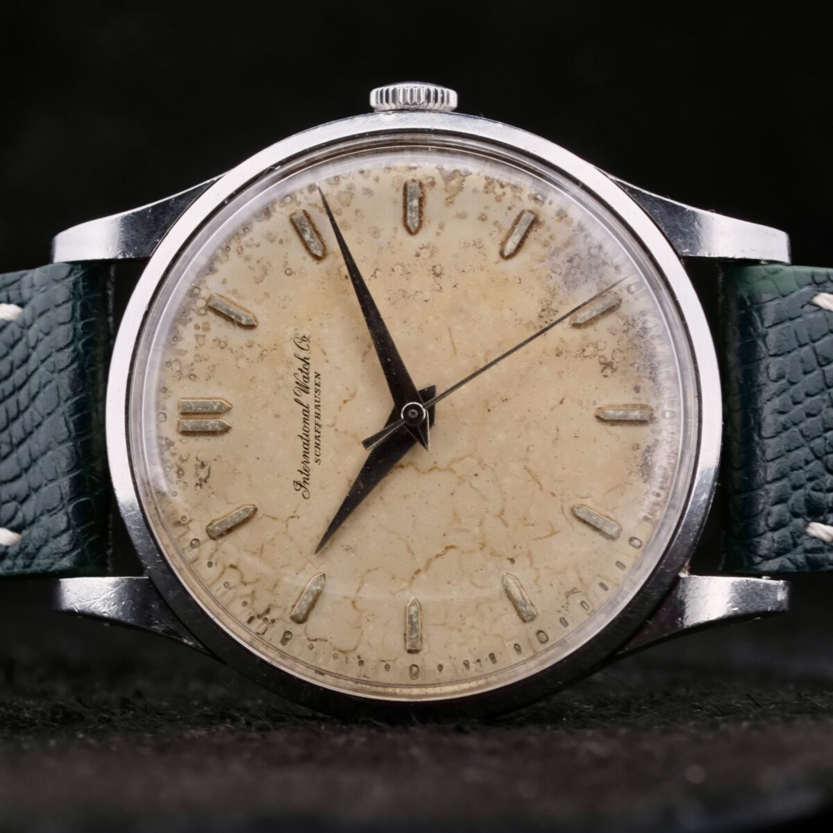 The IWC Caltrava Jumbo Cal. 89 Stainless Steel wristwatch boasts a worn, cream-colored dial with black markers and hands, all encased in metal. Its distressed green leather strap enhances its vintage appeal, while the partially visible brand name on the dial adds timeless elegance.