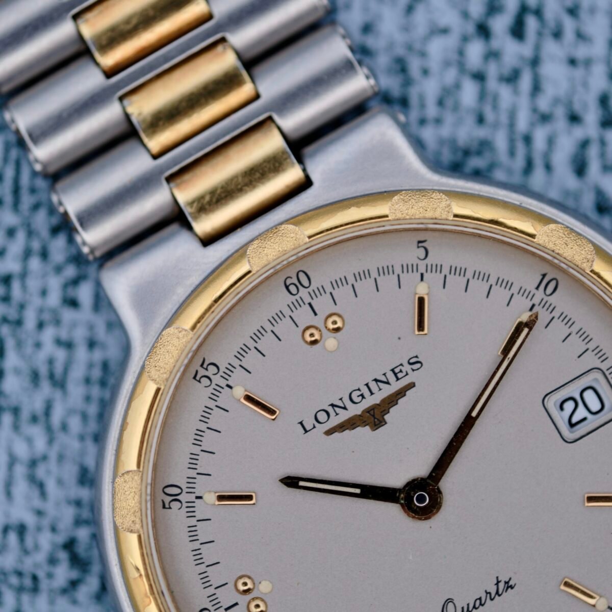 Close-up of a Longines Conquest Quartz Two-Tone 4940 watch on a textured surface, featuring a silver and gold link bracelet, cream dial, gold-tone markers, date window displaying the 20th, black hands, and logo beneath the brand name.